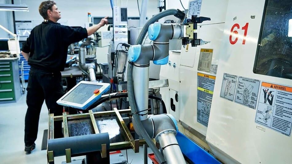 In einem CNC Maschinenpark bestückt ein kollaborierender Roboterarm ohne Schutzkäfig im Vordergrund eine Maschine. Im Hintergrund kontrolliert ein Mensch eine andere Maschine.