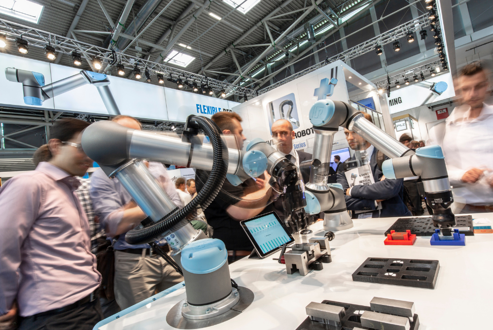 An einem Messestand handhaben zwei Cobots von Universal Robots Bauteile. Im Hintergrund betrachten Menschen die Anwendung und reden miteinander.