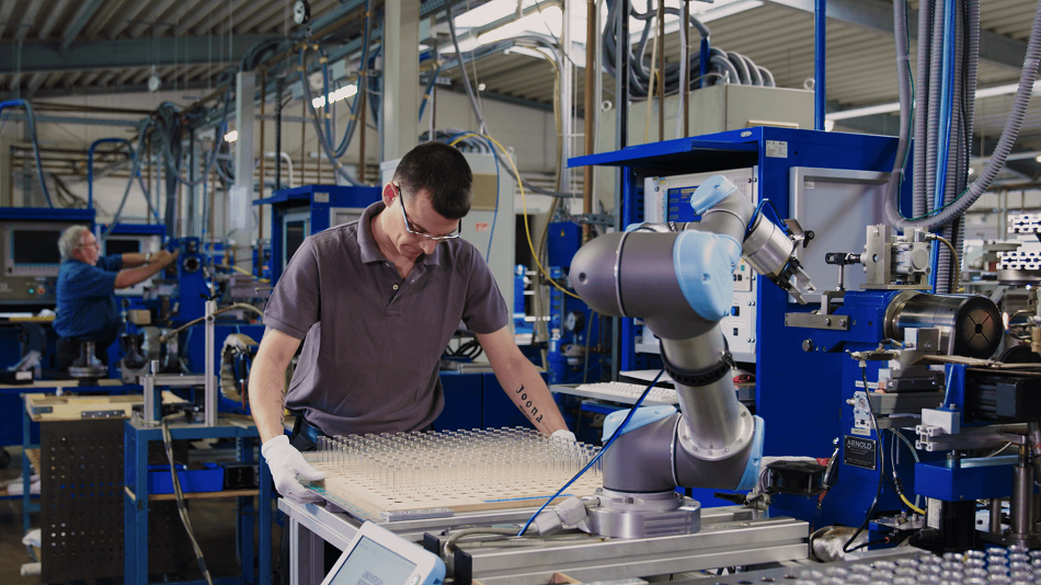Ein Mann stellt einem kollaborierenden Roboterarm ein Tray mit Glasbauteilen bereit, die er zur Bestückung einer Maschine aufnimmt.