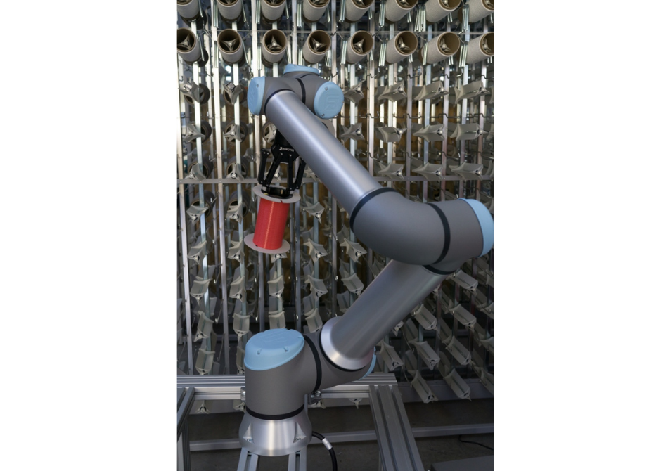 Ein Cobot von Universal Robots bestückt ein Spulengatter mit einer Spule mit rotem Faden.