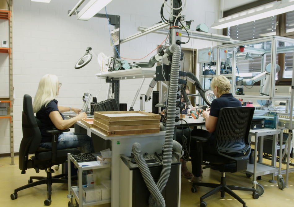 Zwei Frauen sitzen an ihren Arbeitsplätzen in einer Produktion. Rechts neben ihren Tischen steht ein Cobot von Universal Robots in einem Glaskasten.