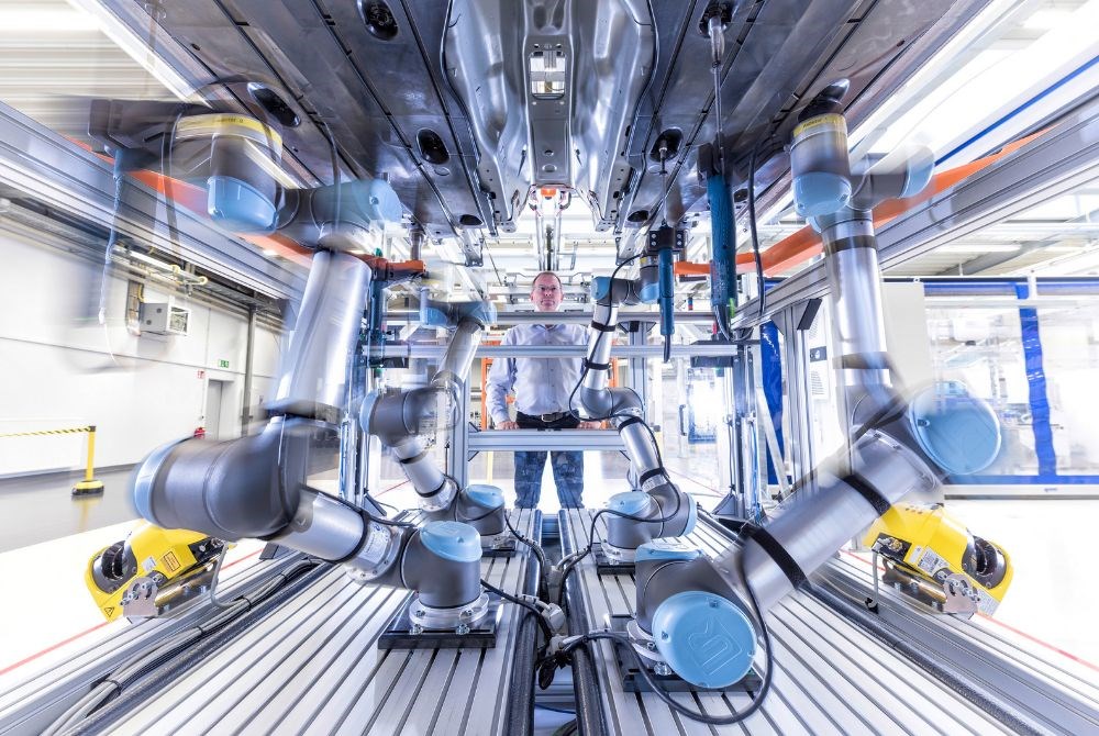 Ein Mann beobachtet stolz vier kollaborierende Roboterarme, die Teile an einer Autokarosse von unten anschrauben.