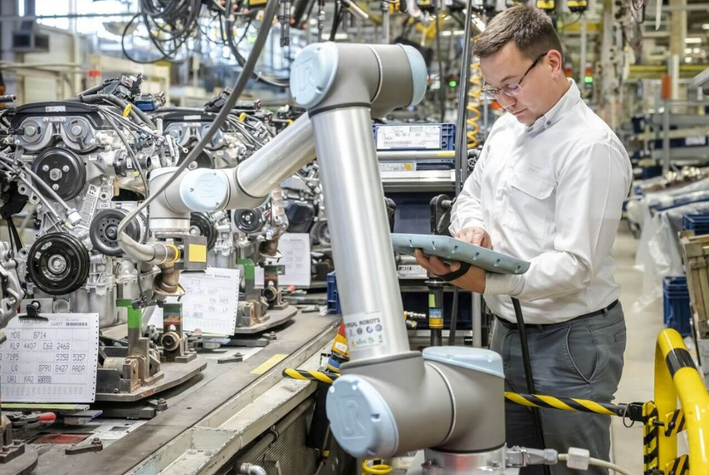 Ein Mann mit Brillen schaut konzentriert auf ein Teach Pendant von einem UR-Roboter. Der Roboterarm montiert ein Autogetriebe an einem Fließband.