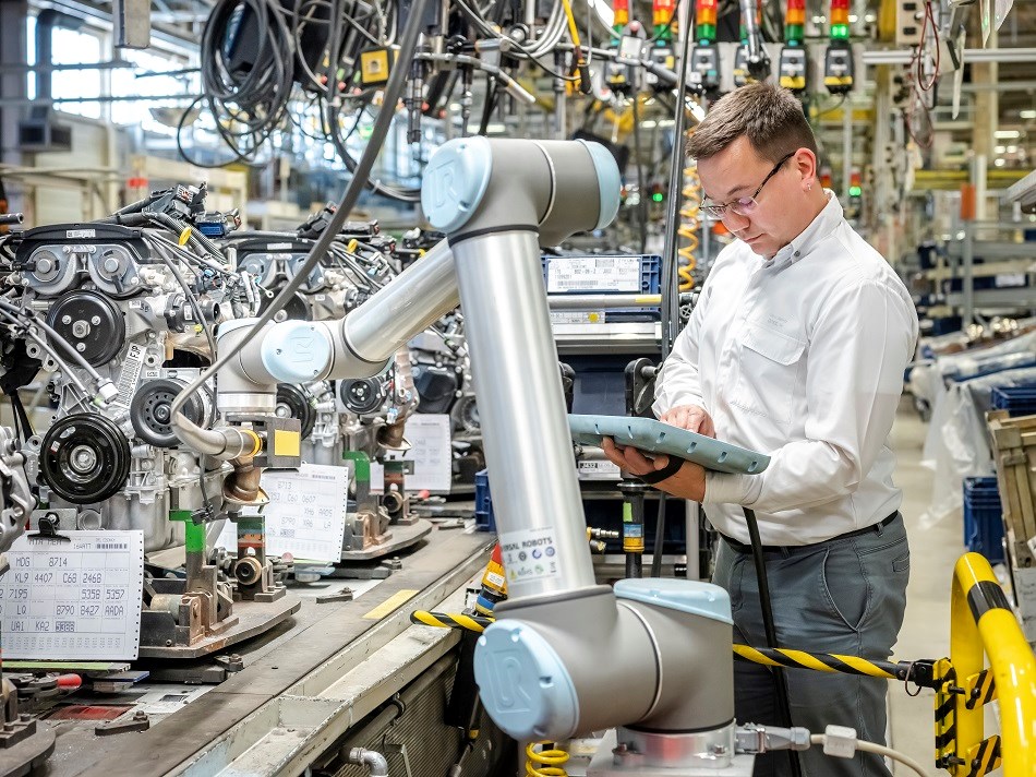 Ein Mann mit Brillen schaut konzentriert auf ein Teach Pendant von einem UR-Roboter. Der Roboterarm montiert ein Autogetriebe an einem Fließband.
