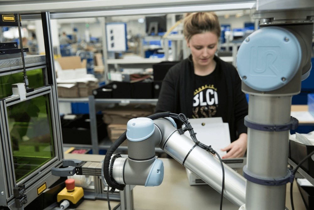 Ein Cobot steht vor einer Lasermaschine. Im Hintergrund ist eine Frau zu sehen, die einen Karton faltet.