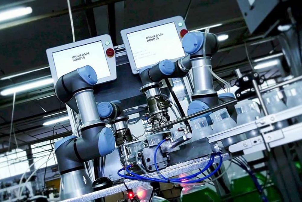 Zwei Cobots von Universal Robots arbeiten an einem Förderband, auf dem Plastikbehälter vorbeilaufen. Im Hintergrund hängen ihre Teach Pendants.