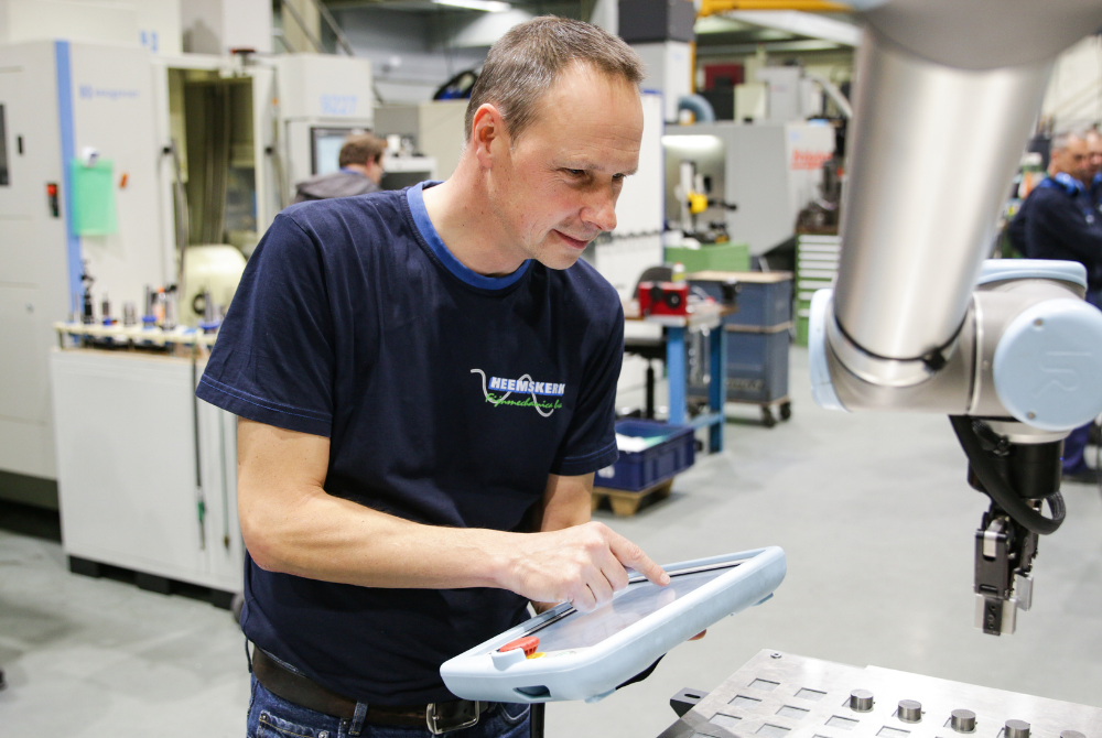 Ein Mann hält das Teach Pendant eines Cobots in der Hand, der im Bildvordergrund zu sehen ist und ein Metallteil greift.