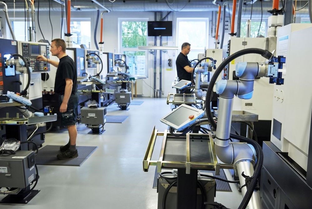 Zwei Männer arbeiten in einem CNC Maschinenpark konzentriert an kollaborierenden Roboterarmen, die die Fräsen mit Bauteilen bestücken.