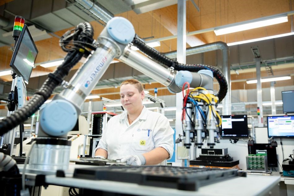 Ein Roboterarm und eine junge Frau arbeiten am selben Arbeitsplatz, an dem Platinen verpackt werden.