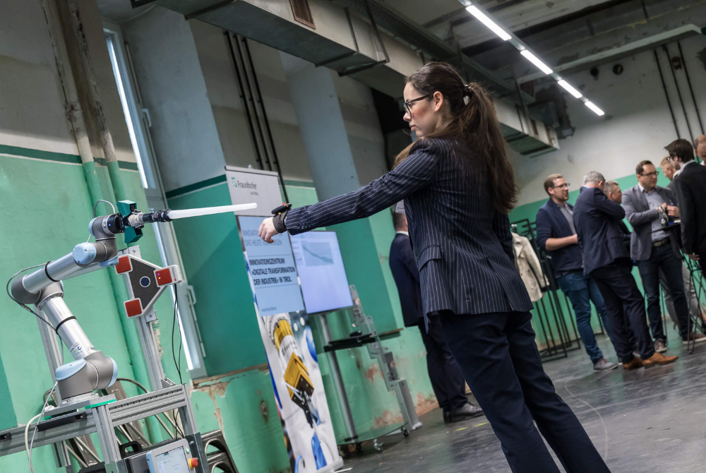 Eine junge Frau mit einem Sensor an ihrem linken Handgelenk steht vor einem Roboterarm, der ein Messgerät an den Sensor hält.