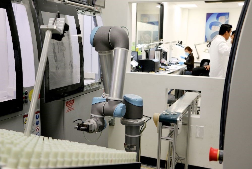 Ein kollaborierender Roboterarm bestückt eine Maschine in einem Labor. Ein Fließband führt in einen Raum hinter eine Scheibe, wo Menschen arbeiten.