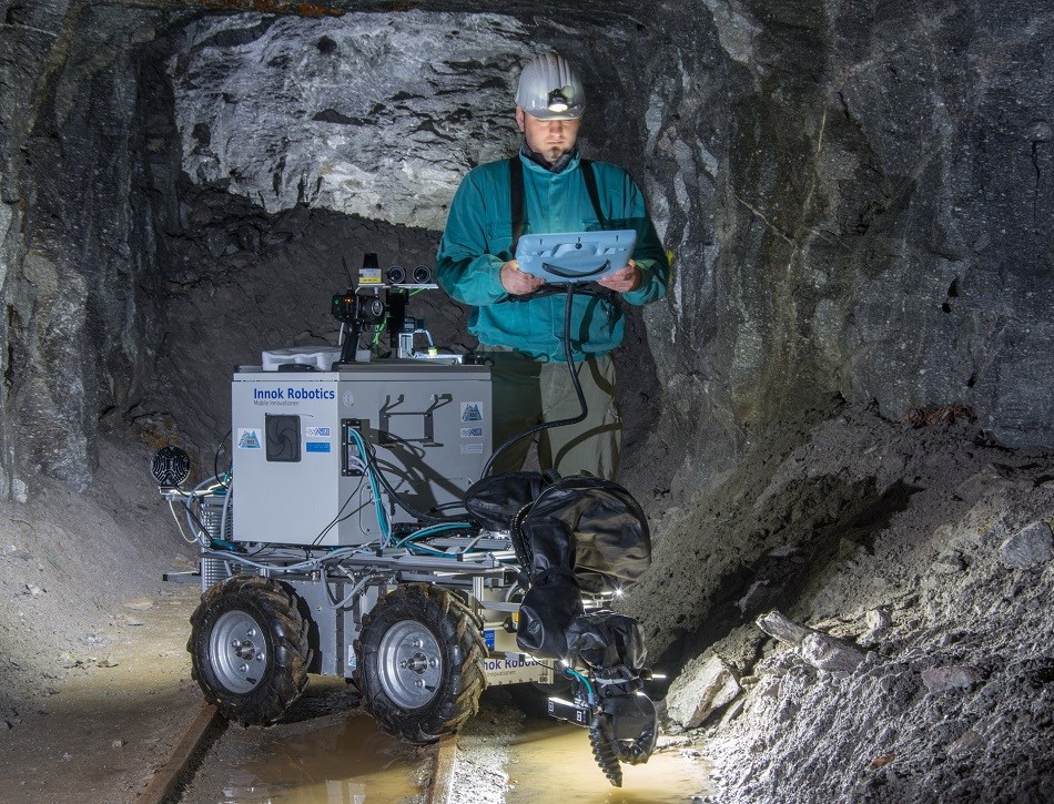 Ein Mann hält ein Bedienpanel in der Hand und programmiert damit einen kollaborierenden Roboterarm auf einem mobilen Wagen in einem Bergwerk.
