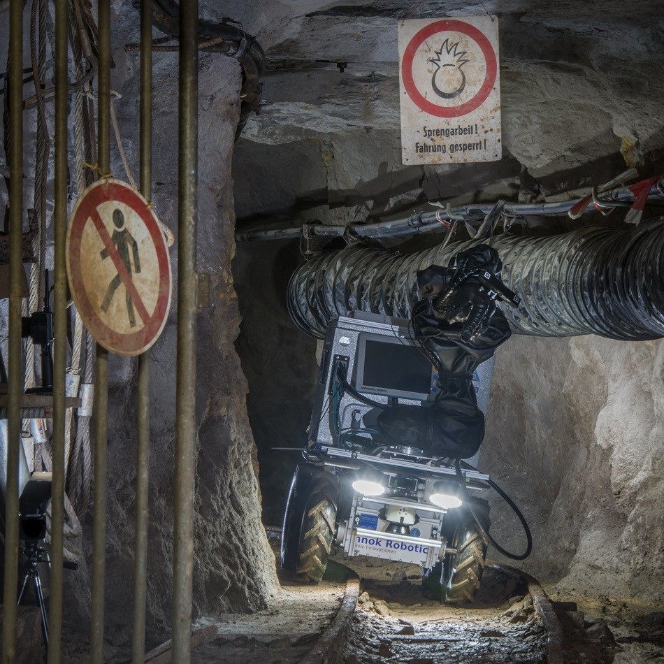 Ein kollaborierender Roboter arbeitet an einem gefährlichen Bergwerkgebiet. Warnschilder sagen aus, dass Menschen hier nicht erlaubt sind und der Weg wegen Sprengarbeiten gesperrt ist.