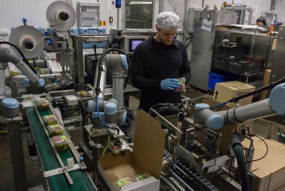 Ein Mann und drei kollaborierende Roboterarme verpacken zusammen Gütern an einem Laufband in Kartons.