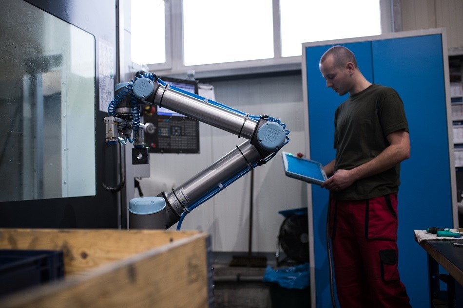Ein Mann bedient einen kollaborierenden Roboterarm, der ohne Schutzumhausung an einer Werkzeugmaschine arbeitet.