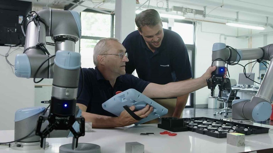 Ein älterer Mann hält ein Bedienpanel und berührt einen Roboterarm mit Greifer. Neben ihm schaut ein junger Mann lächelnd zu. Im Vordergrund steht ein zweiter Cobot mit Greifer.