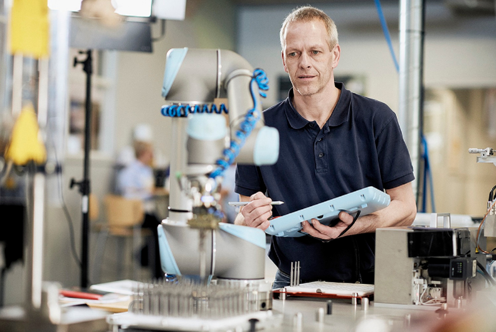 Ein Mann, der ein Control Panel in seiner Hand hält, beobachtet konzentriert einen Roboterarm. In seiner rechten Hand hält er einen Stift. Der Roboterarm greift Metallteile.