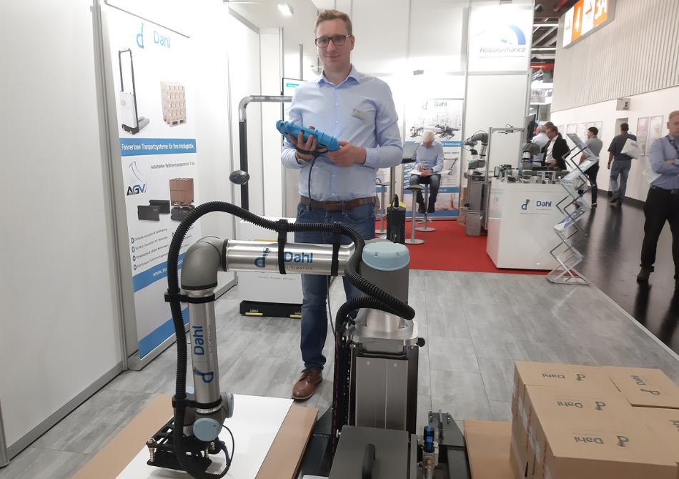 Ein junger Mann steht mit einem Control Panel hinter einem kollaborierenden Roboterarm. Der Cobot stapelt Kisten.