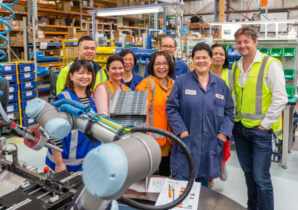 Ein Team aus neun Kollegen stehen lächelnd in einem Industriebetrieb und posiert hinter einem kollaborierenden Roboterarm.