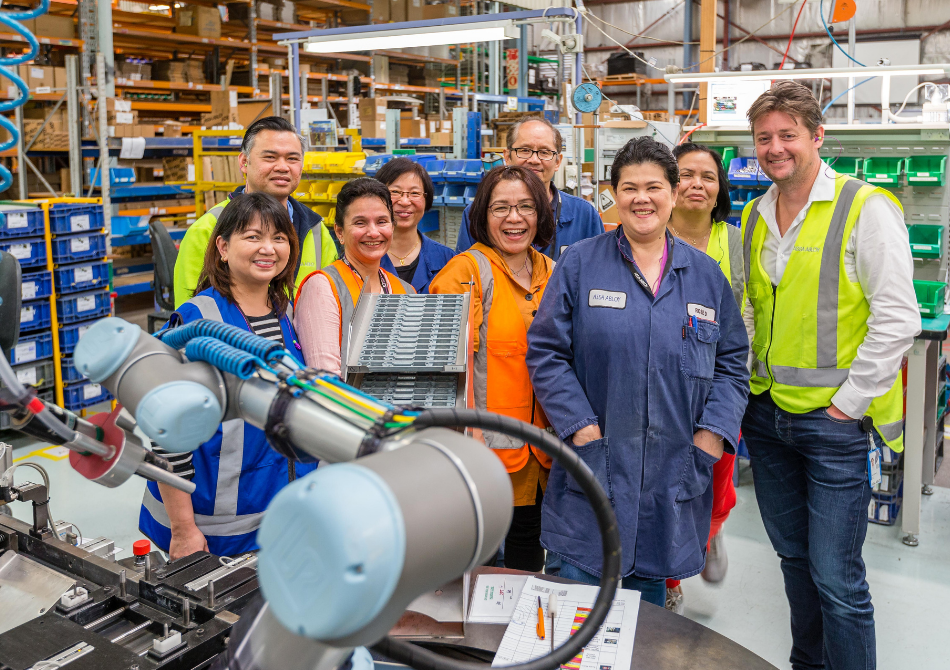 Ein Team aus neun Kollegen stehen lächelnd in einem Industriebetrieb und posieren hinter einem kollaborierenden Roboterarm.