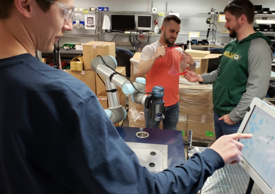 Zwei junge Männer kontrollieren eine Gesichtsmaske. Sie stehen neben einem Cobot. Ein anderer junger Mann im Vordergrund wählt eine Taste auf einem Teach Pendant des Roboters aus.