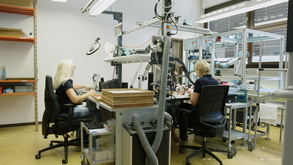 Zwei Frauen sitzen an ihren Tischen in einer Elektronikfertigung und arbeiten mit Roboterarmen zusammen.