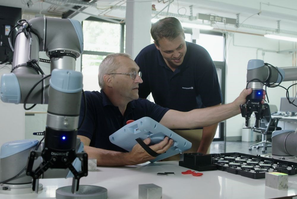 Ein älterer Mann hält ein Bedienpanel und berührt einen Roboterarm mit Greifer. Neben ihm schaut ein junger Mann lächelnd zu. Im Vordergrund steht ein zweiter Cobot mit Greifer.