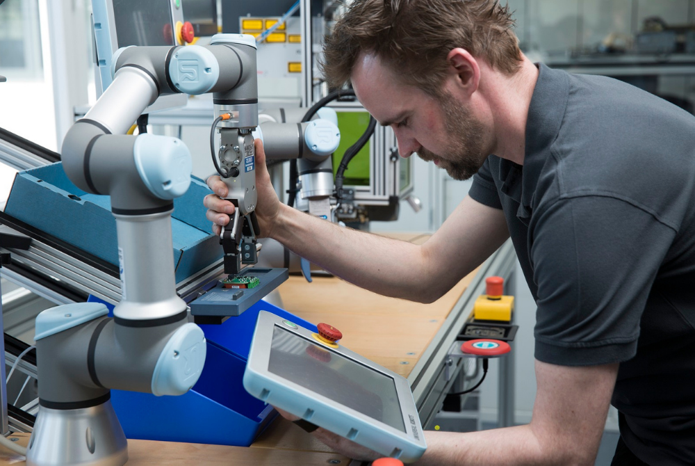 Ein junger Mann führt mit seiner Hand einen Cobot zu einem Elektronikbauteil. Er schaut konzentriert auf das Teach Pendant des Roboters.