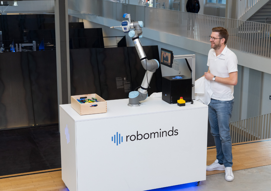 Ein junger Mann steht neben einem Stand mit einem Roboterarm und einer Kiste, die mit Süßigkeiten gefüllt ist.