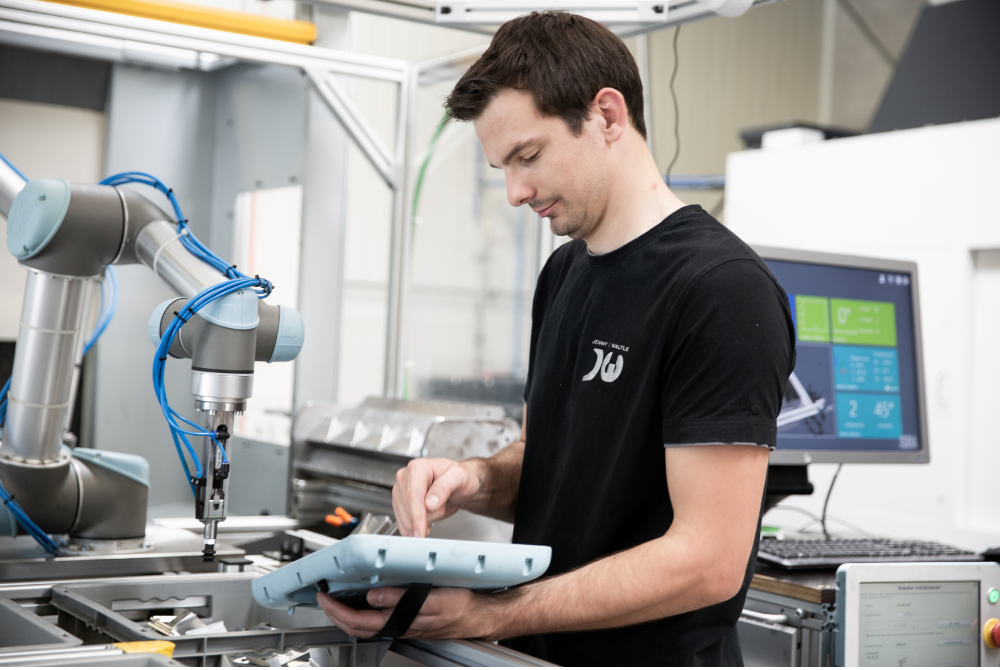 Ein junger Mann hält das Teach Pendant eines Cobots von Universal Robots in der Hand und programmiert darauf etwas. Der Roboterarm steht neben ihm an einer CNC-Fräse.