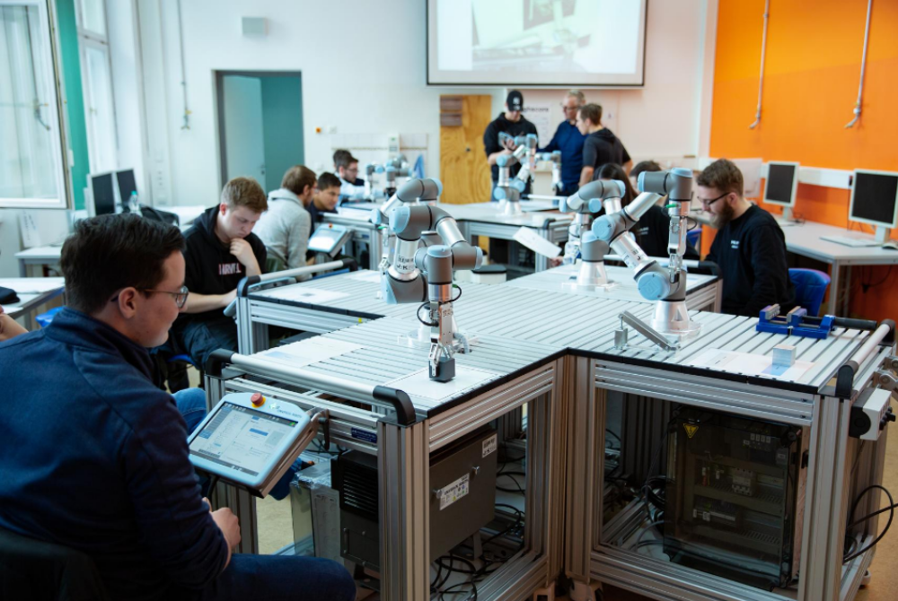 Eine Klasse mit 12 Schülern sitzen an Tischen, auf denen Cobots angebraucht sind. Sie lernen den Umgang mit den Roboterarmen.