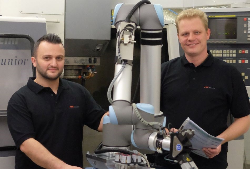 Zwei Männer stehen neben einem Roboterarm. Im Hintergrund ist eine CNC Fräsmaschine zu sehen. Der Mann links vom Roboterarm berührt diesen. Der Mann rechts vom Roboterarm hält ein Heft in der Hand und lächelt in die Kamera.