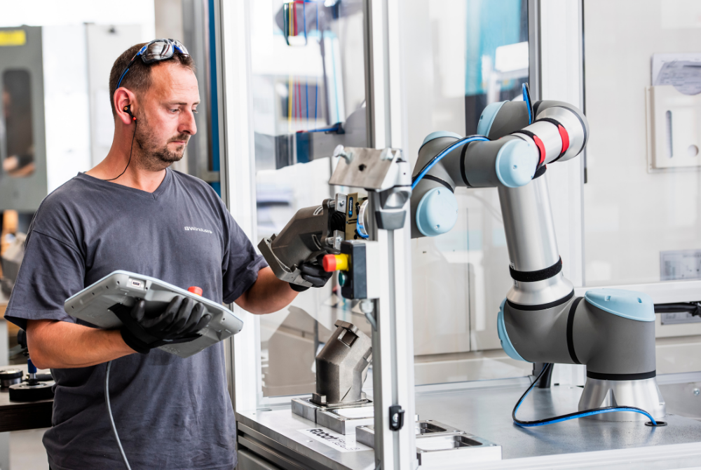 Ein Werker mit Schutzbrille auf der Stirn hält das Teach Pendant eines UR-Cobots in der Hand und greift nach einem Metallteil, dass der Cobot gefasst hält.