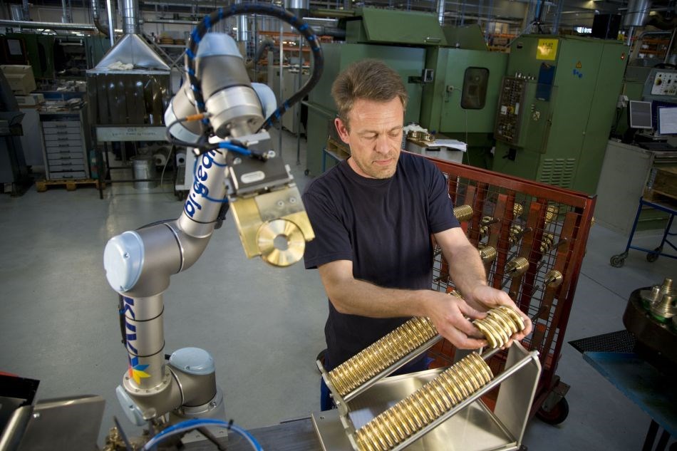 Ein Cobots hält ein goldenes Werkstück, während ein menschlicher Werker ihm diese Werkteile in einem Register zur Verfügung stellt und einordnet.
