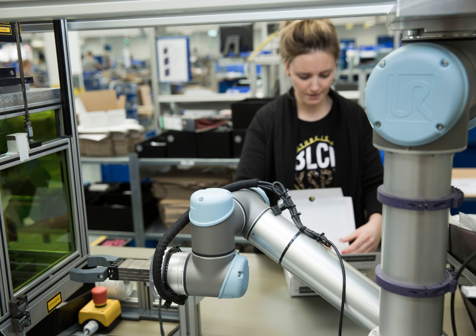 Ein Roboterarm wartet vor einer Lasermaschine mit einem Greifer. Im Hintergrund ist unscharf eine dunkelblonde Frau zu erkennen, die einen Karton faltet.