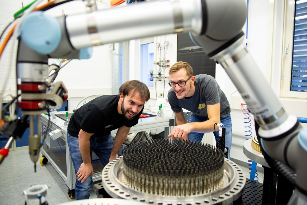 Zwei Mitarbeiter der ArianeGroup schauen auf einen Roboterarm, der Prüfarbeiten an einem Raketentriebwerk vornimmt.