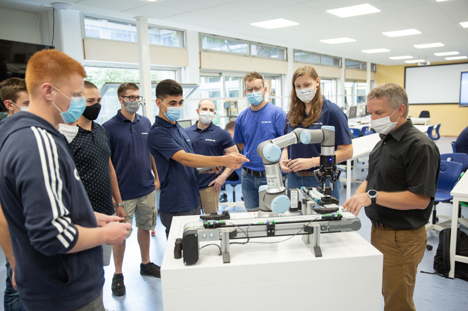 Hier sieht man eine Klasse mit Schülern und einen Lehrer, ein Schuler zeigt auf den Cobot auf und erzählt was. Alle hören zu.