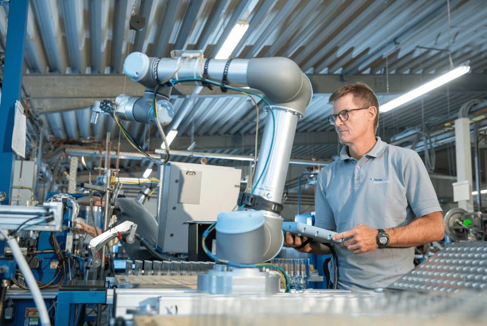 Ein Roboter ist links im Bild zu sehen. Rechts davon steht ein Mann, der den Roboter programmiert über ein Teach Pendant von Universal Robots.