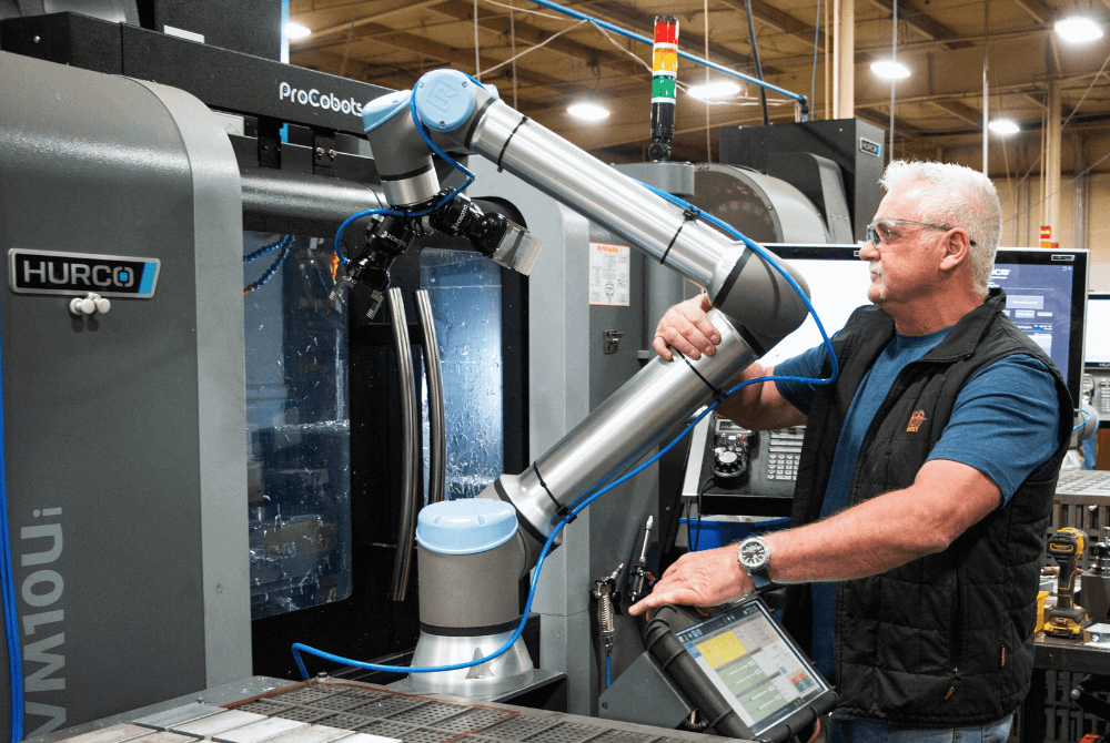 Links im Bild steht eine CNC-Fräse mit dem Aufdruck "Hurco". Davor steht ein Roboterarm mit blauen Kappen, der an seinem mittleren Teil von einem Mann mit der rechten Hand gegriffen wird. Der Mann hat weiße Haare, einen Bart, trägt ein blaues T-Shirt und eine schwarze Weste.