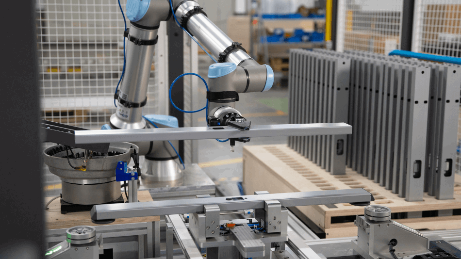 Ein Roboterarm hält einen Tischfuß-Ausleger und legt ihn in eine Vorrichtung.