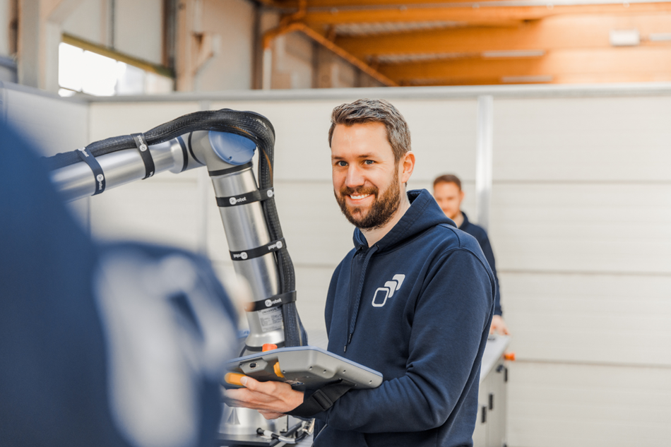 Hier sieht man einen Mitarbeiter aus moduco GmbH lächelend neben einen Roboter. 