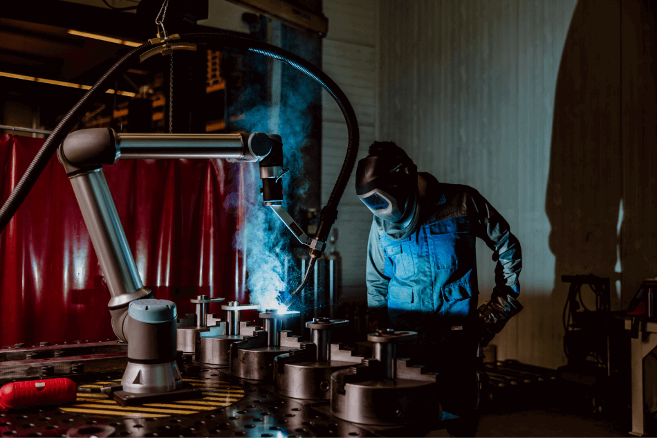 Ein Cobot schweißt ein Metallteil. Neben ihm steht ein Mensch mit Schweißmaske.