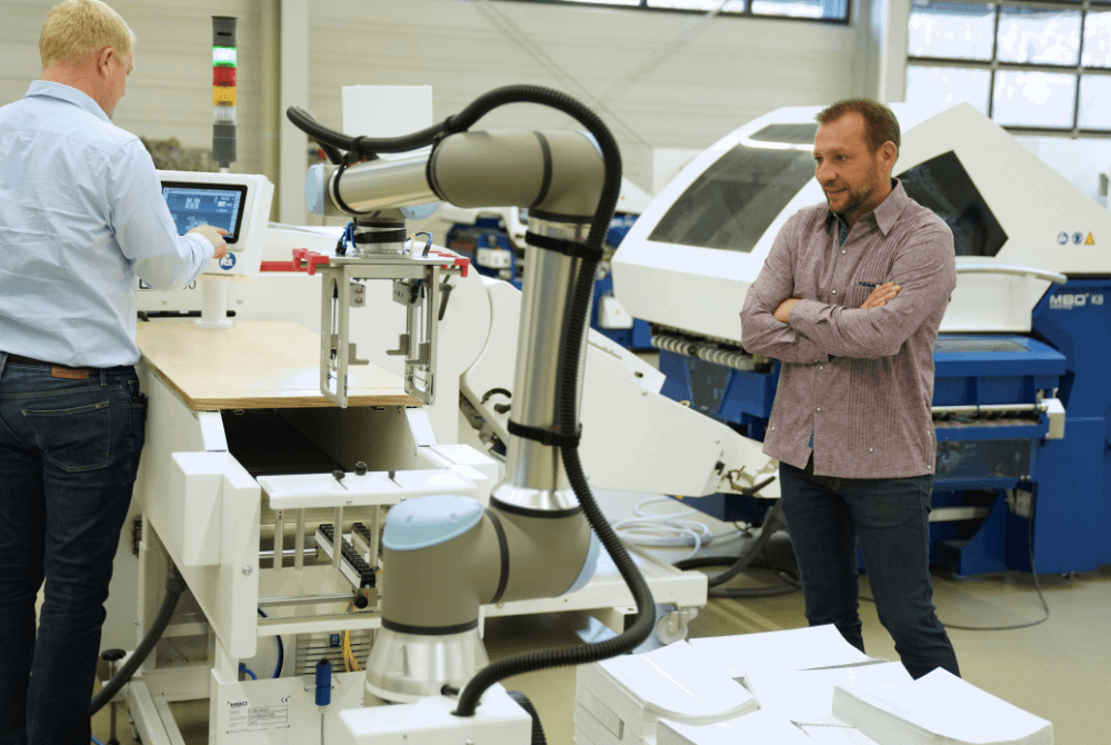 Eine Ansicht auf ein Druckerei mit Falzmaschinen. Im vorderen Bildbereich ist ein kollaborierender Roboterarm zu sehen, der Papier abstapelt. Neben ihm steht ein Werker.
